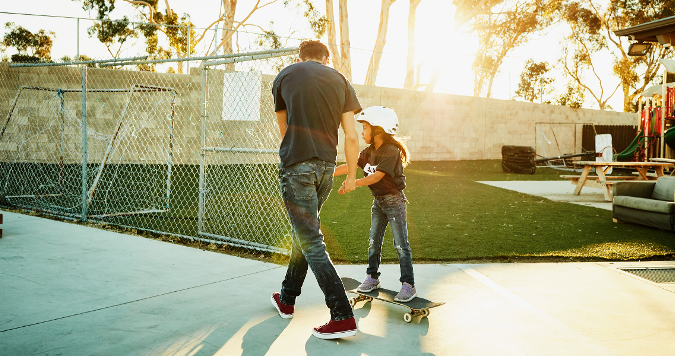 A dad helping his daughter learn to skateboard at the park at golden hour